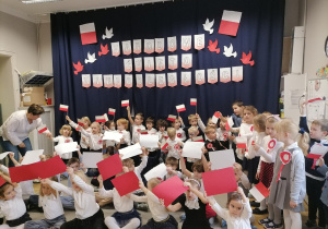 Grupa dzieci w strojach galowych tańcząca z białymi i czerwonymi kartkami.