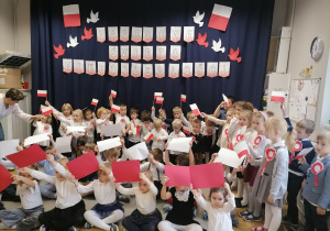 Grupa dzieci w strojach galowych tańcząca z białymi i czerwonymi kartkami.