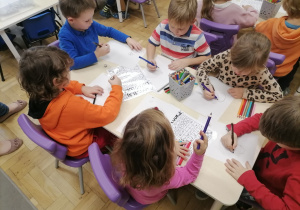 Grupa dzieci siedząca przy stoliku i rzucająca kośćmi do gry rysując liścioludka.