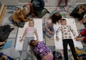 Chłopiec i dziewczynka leżą na białych kartonach a pozostałe dzieci ich odrysowują