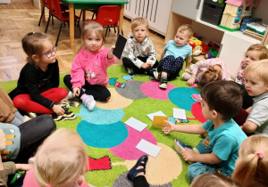Dzieci siedzą na dywanie a jedna dziewczynka pokazuje wylosowaną czarną kartkę