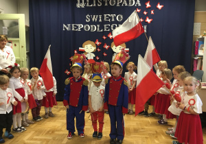 Na pierwszym planie stoją dwaj chłopcy ubrani w ułańskie mundury oraz dziewczynka w biało- czerwonym stroju.