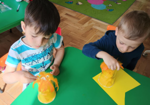 Dwóch chłopców przykleja żółte piórka do plastikowego kubeczka