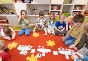 Grupa dzieci siedzi na dywanie i układa zadania matematyczne związane z pączkami.
