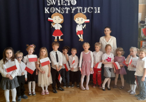 Grupa dzieci wraz z nauczycielka prezentuja wiersz patriotyczny na tle dekoracji z okazji Konstytucji 3 Maja