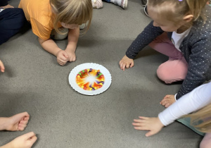 Siedzące dzieci na dywanie i obserwujące tworzącą się tęczę na talerzu z kolorowych cukierków.