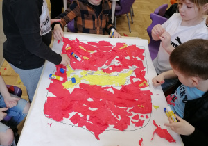 Grupa dzieci wykleja herb Łodzi bibułą w kolorze czerwonym i żółtym.