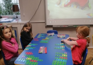 Dzieci siedzą przy stole, w tle na projektorze widać filmik instruktażowy odnośnie wykorzystania klocków NEMI