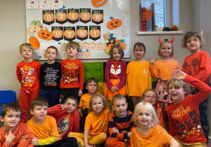 Grupa usmiechniętych dzieci ubranych na pomarańczowo pozuje do zdjęcia na tle dyniowej dekoracji.