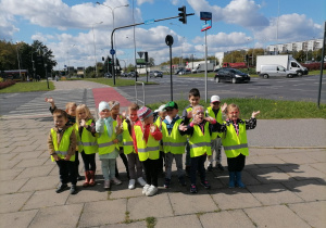Grupa dzieci w odblaskowych kamizelkach pozuje do zdjęcia na tle skrzyżowania