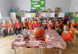 Grupa dzieci w pomarańczowych strojach pozuje do zdjęcia z wielką dynią