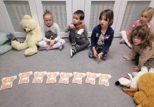 Dzieci siedzą na dywanie, przed nimi leżą misie z numerami.