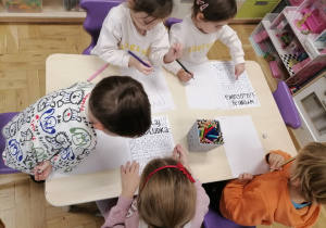 Grupa dzieci siedząca przy stoliku i rzucająca kośćmi do gry rysując liścioludka.