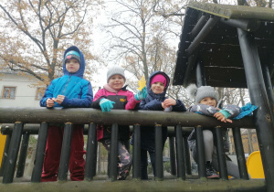 Dzieci w ciepłych kurtkach pozują do zdjęcia stojąc na drewnianym mostku na placu zabaw