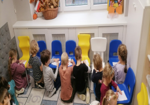 Dzieci siedzą na dywanie przy krzesełkach i rysują