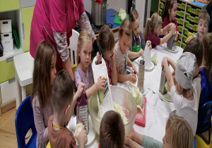 Dzieci wraz z nauczycielką oglądają warzywa i przybory kuchenne znajdujące się na stole