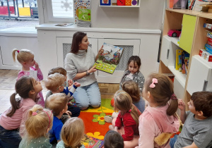 Nauczycielka prezentuje dzieciom książkę o dinozaurach, dzieci siedzą w kole na dywanie.