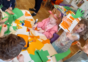 Grupa dzieci przy przy stoliku wykonuje marchewki z pomarańczowych i zielonych pasków.