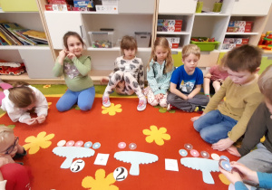 Grupa dzieci siedzi na dywanie i układa zadania matematyczne związane z pączkami.