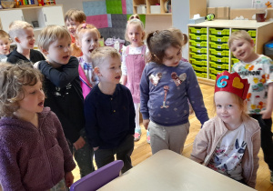 Grupa dzieci stoi przy stoliku przy którym znajduje się dziewczynka, która obchodzi urodziny z czerwoną korona na głowie oraz cyfrą 6.