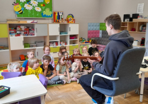 Dzieci siedza na dywanie, przed nimi uczeń Szkoły Podstawowej prezentuje techniki gry na gitarze.