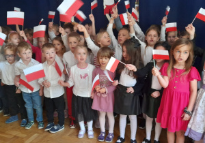 Grupa dzieci na tle granatowej dekoracji podnosi w górę wykonane własnoręcznie flagi Polski.
