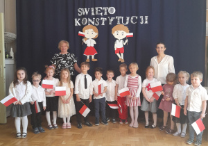 Grupa dzieci wraz z Panią Dyrektor oraz nauczycielką stoja na tle dekoracji z okazji Konstytucji 3 Maja