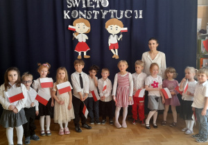 Grupa dzieci wraz z nauczycielka prezentuja wiersz patriotyczny na tle dekoracji z okazji Konstytucji 3 Maja